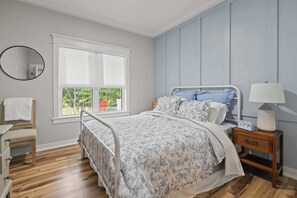 Ein Schlafzimmer mit einem Bett, einem Nachttisch, einem Sessel, einem Spiegel und einem Fenster mit Jalousien.