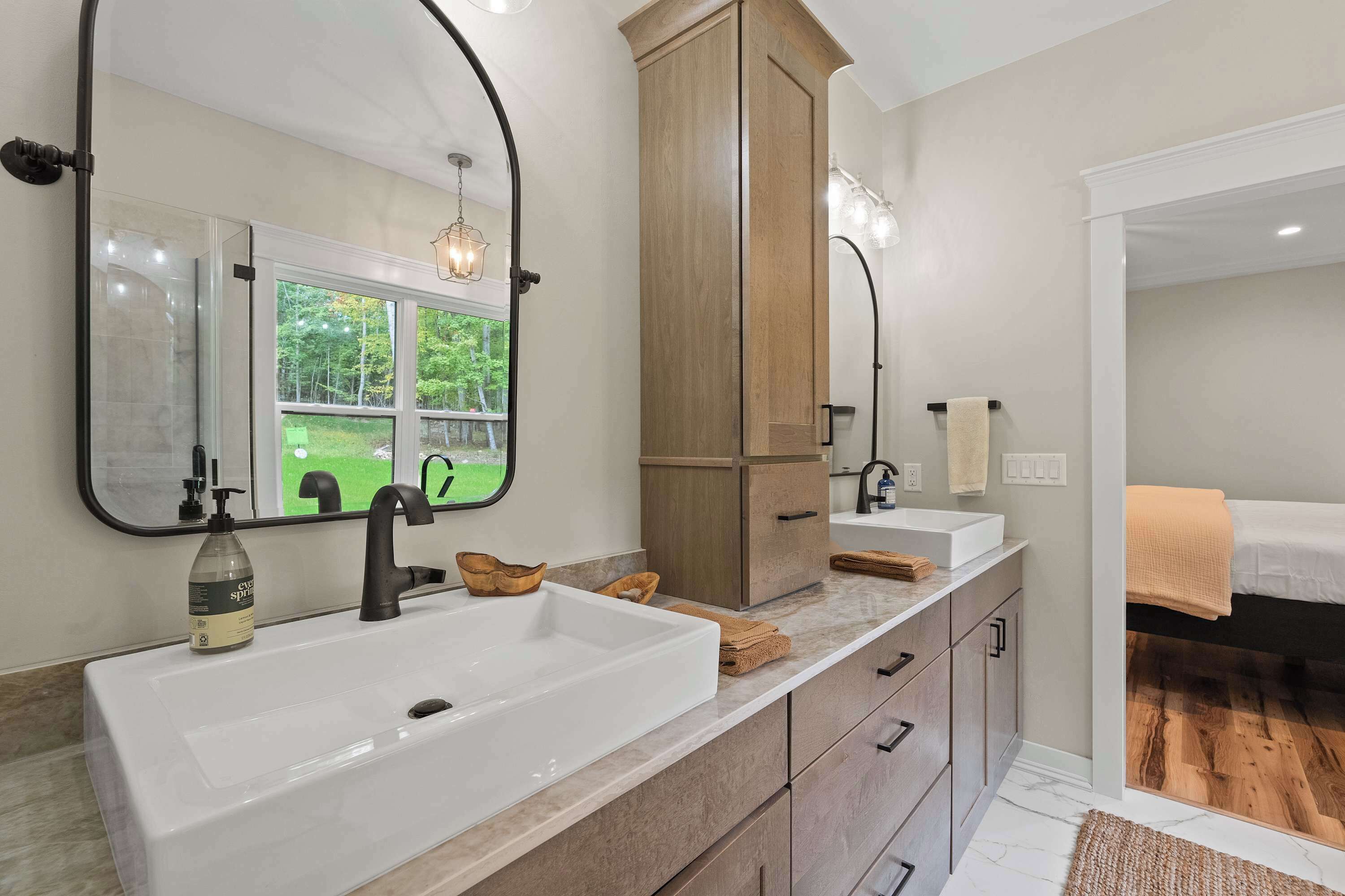 Une salle de bain avec un grand lavabo blanc, des meubles en bois, un miroir et une fenêtre donnant sur un paysage verdoyant.
