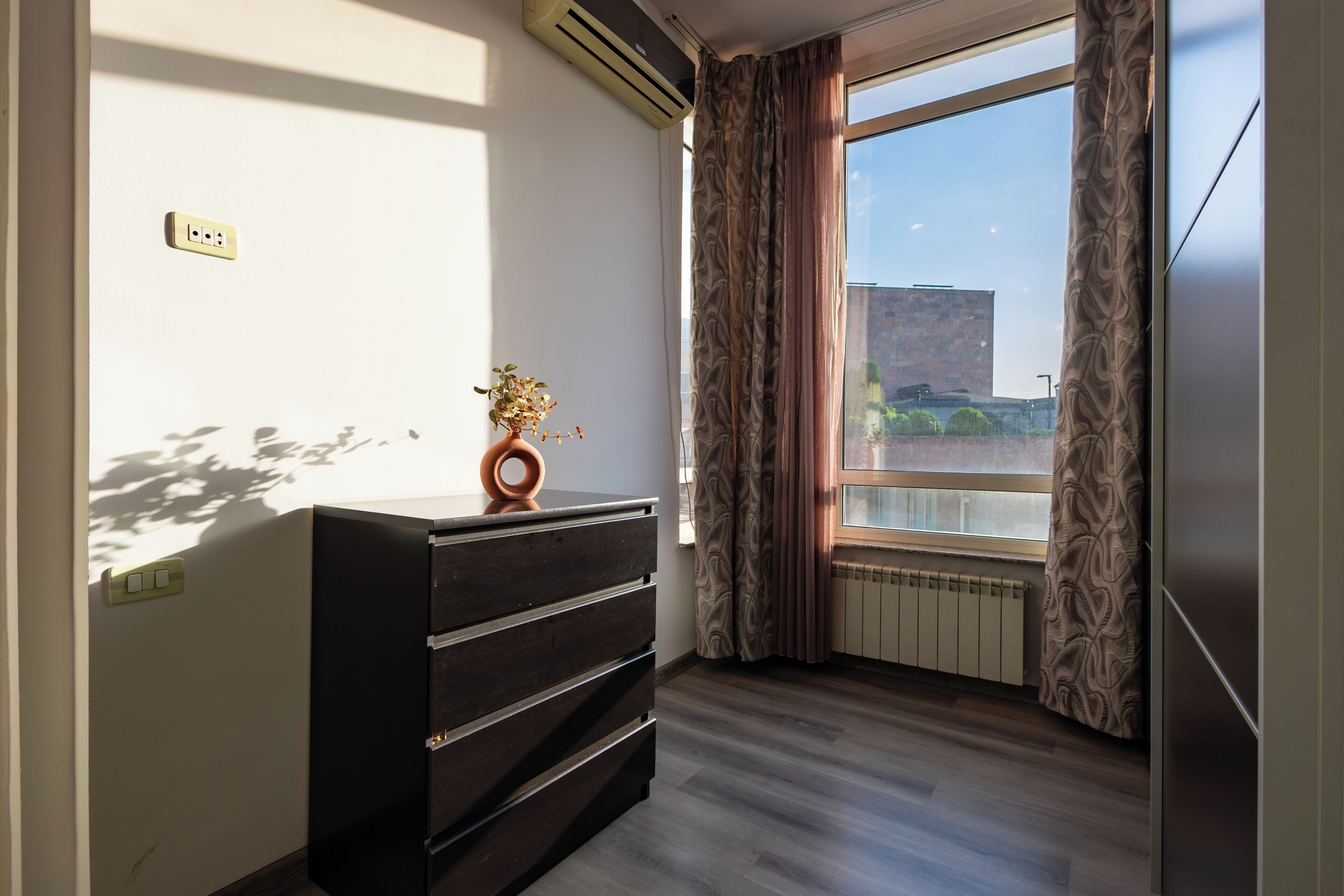 A room with a wooden dresser, a potted plant, and a large window with curtains.