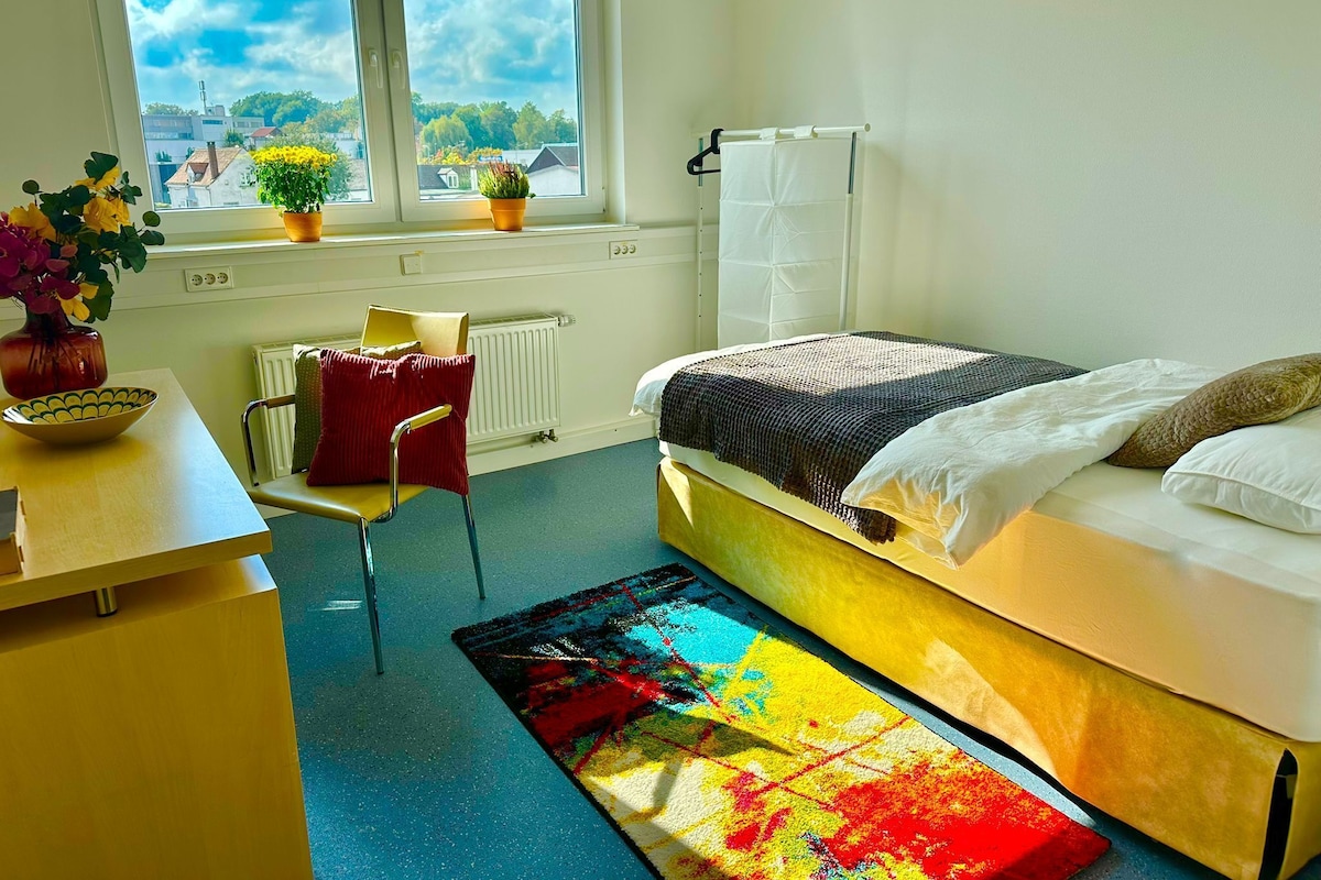 Una camera da letto con un letto, una sedia, un tavolo con una ciotola, un tappeto colorato e una finestra che si affaccia su edifici e alberi.