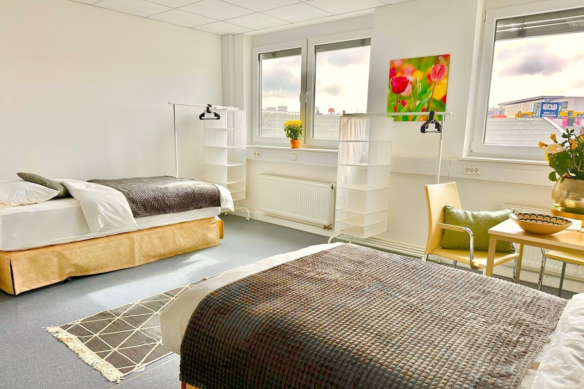 Una camera d'hotel moderna con un letto, una sedia, un tavolino, un termosifone e una finestra con vista sulla città.