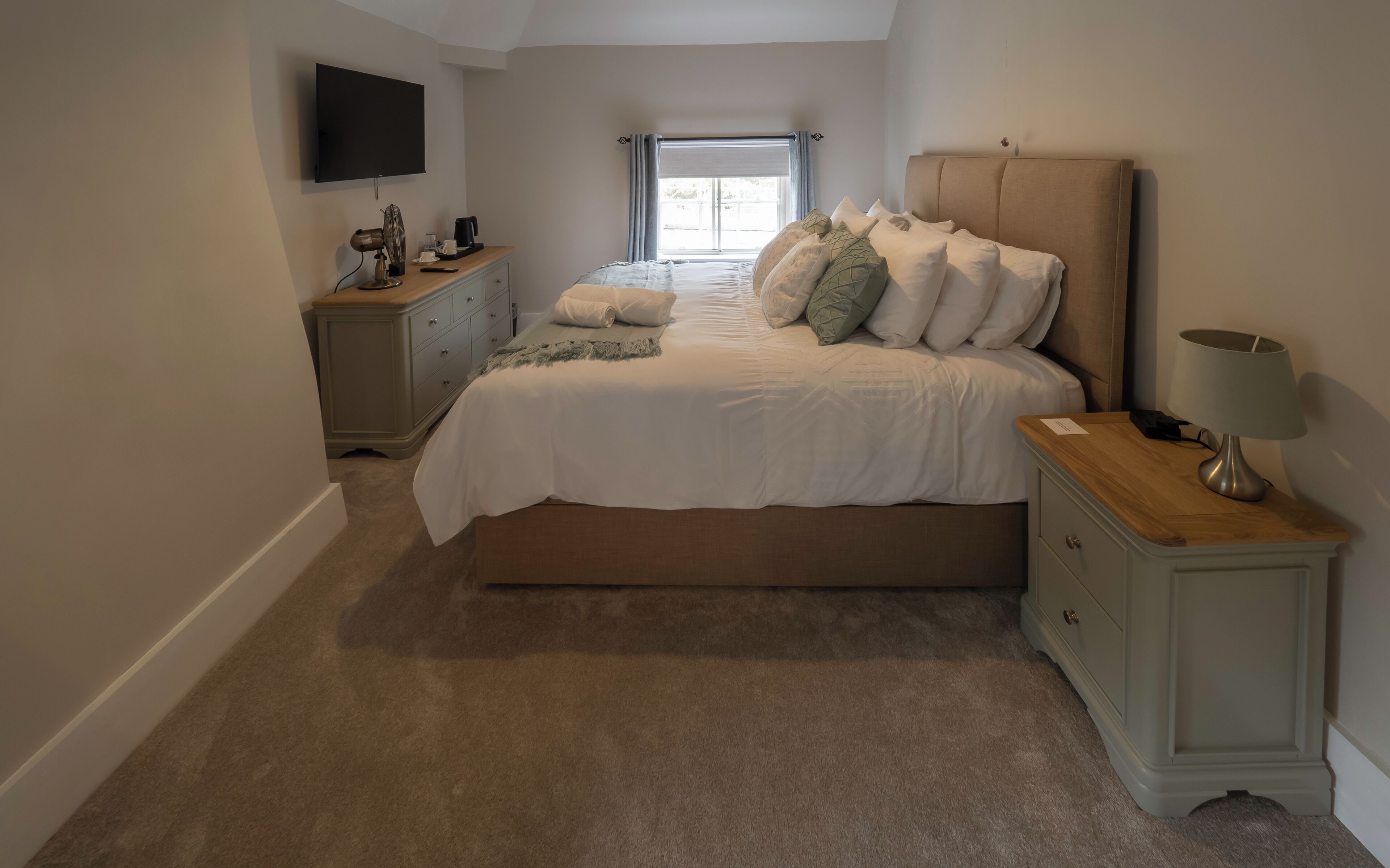 A bedroom with a large bed, a bedside table with a lamp, a dresser, a TV mounted on the wall, and a window with curtains.