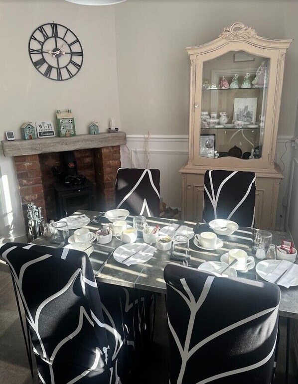 Un comedor con una mesa redonda para cuatro, sillas negras con diseños blancos, una chimenea, un espejo decorativo y un reloj de pared.