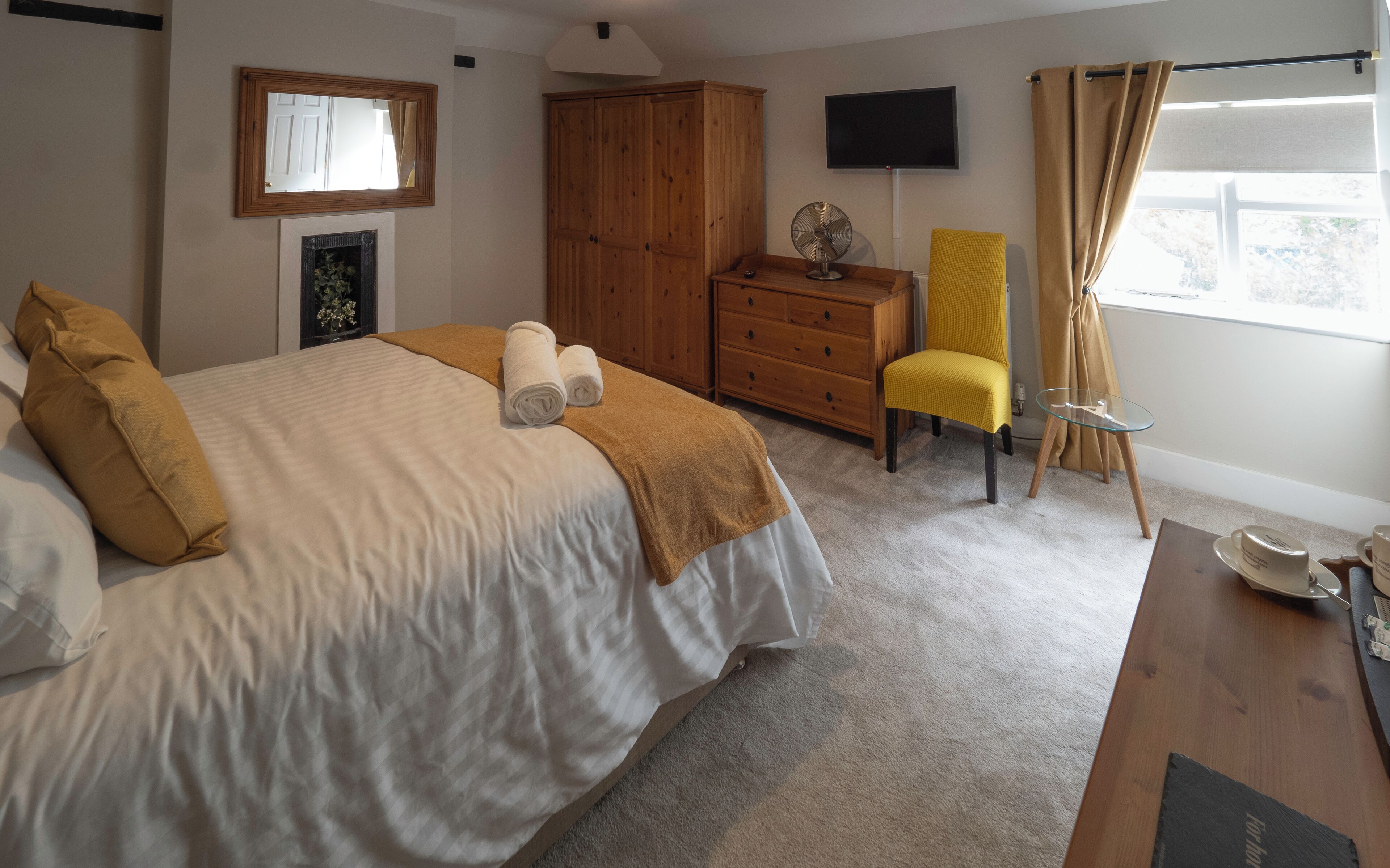Ein Schlafzimmer mit einem Bett, einem Holzschrank, einem Fernseher, einem Ventilator, einem gelben Sessel, einem Glastisch und einem Fenster mit Vorhängen.