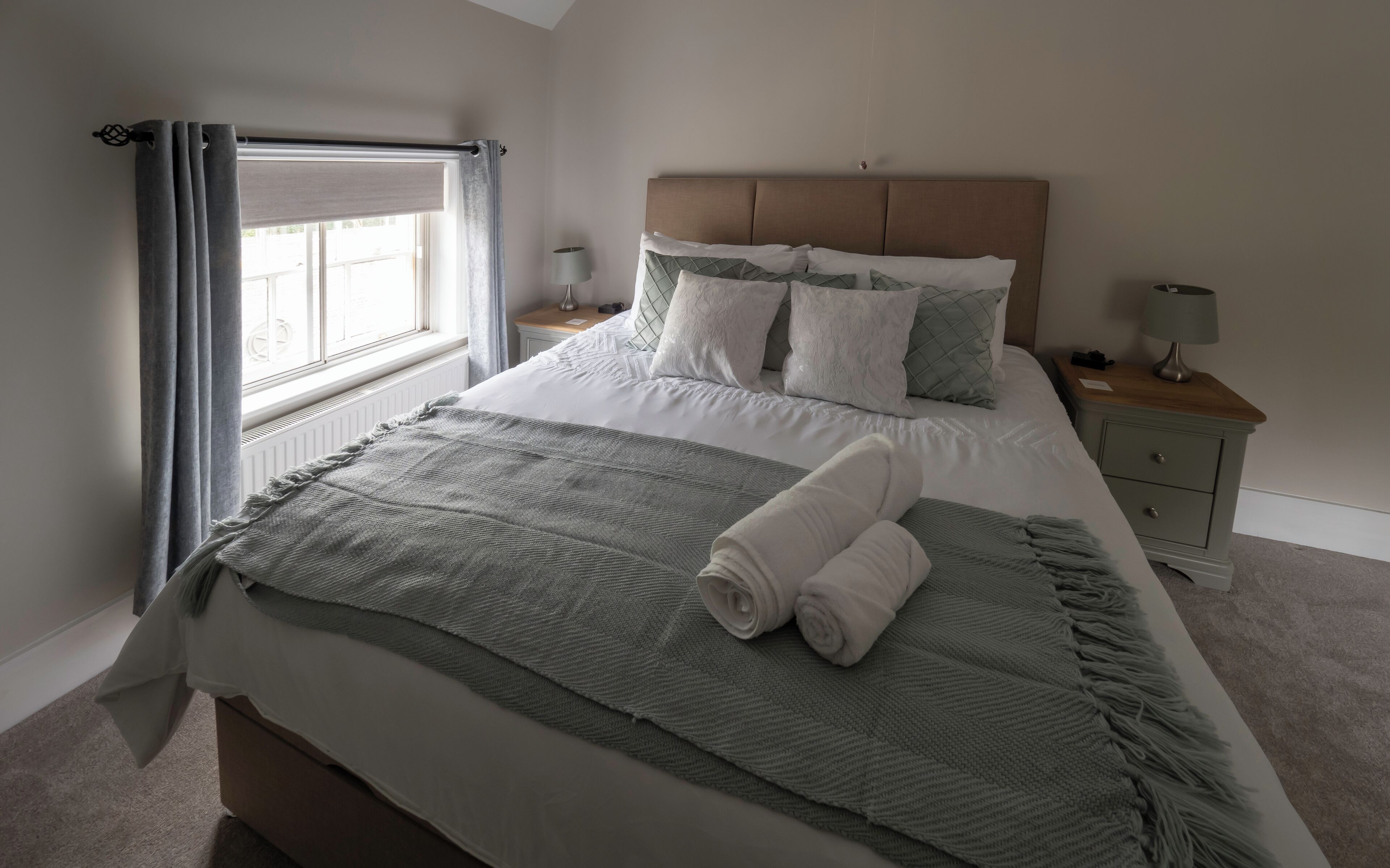 Una cama bien hecha con una manta gris y toallas enrolladas, una lámpara de noche y una mesita de noche con lámpara.