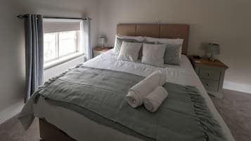 Una cama bien hecha con una manta gris y toallas enrolladas, una lámpara de noche y una mesita de noche con lámpara.