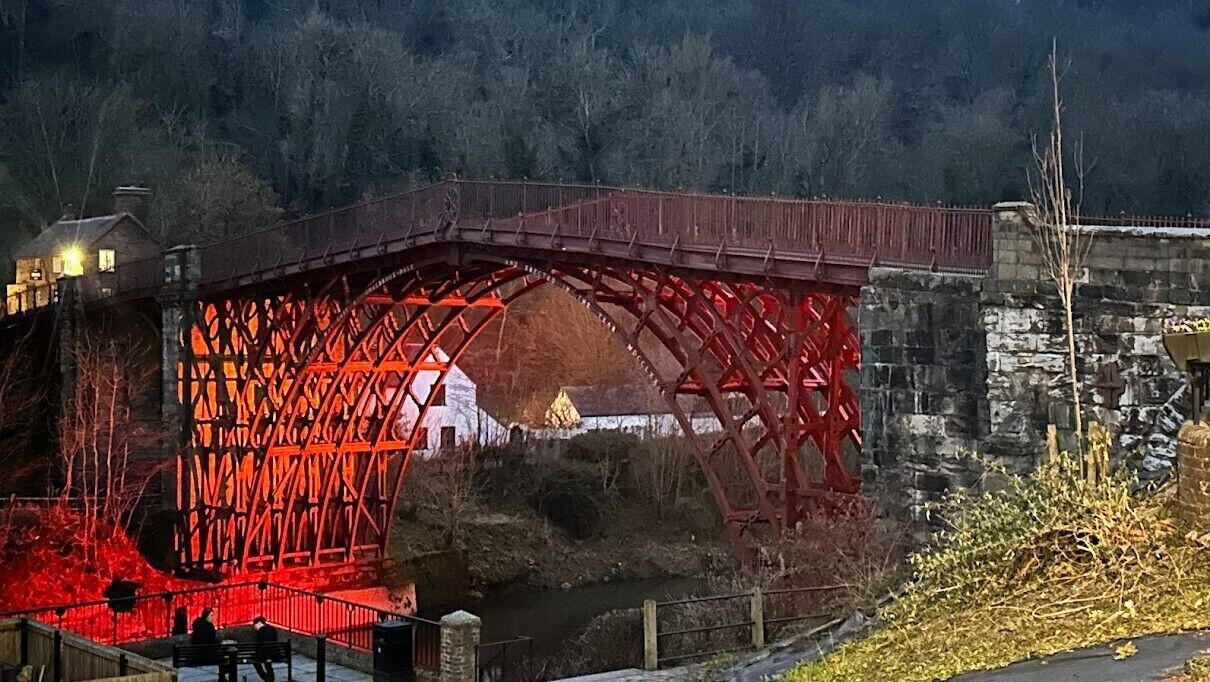 A red metal bridge with a wooden walkway and a stone wall in the background.