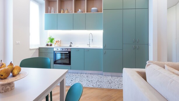 Una cocina moderna con armarios azul verdoso, encimera blanca y una zona de comedor con mesa y sillas.