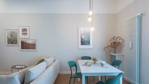 Una sala de estar moderna con un sofá blanco, sillas color turquesa y una mesa de comedor con mantel blanco. Hay cuadros en la pared y un radiador a la derecha.