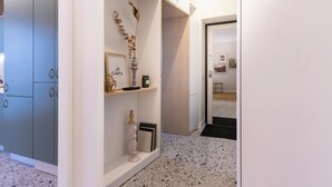 Un pasillo con pared blanca, un armario gris, una estantería con adornos y una puerta que da a otra habitación.