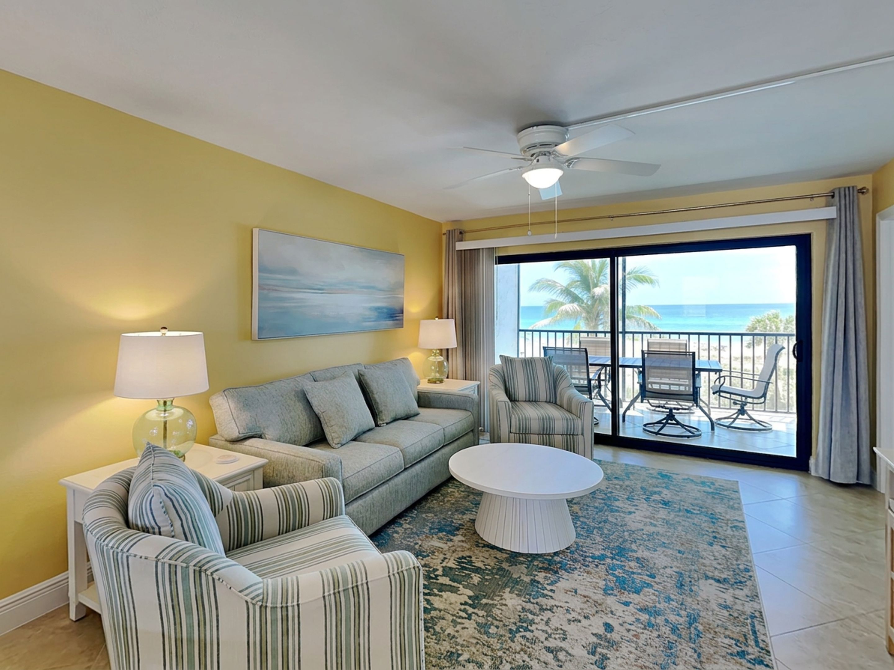 A room with a sofa, armchairs, a coffee table, a ceiling fan, and a view of the ocean through a sliding glass door.