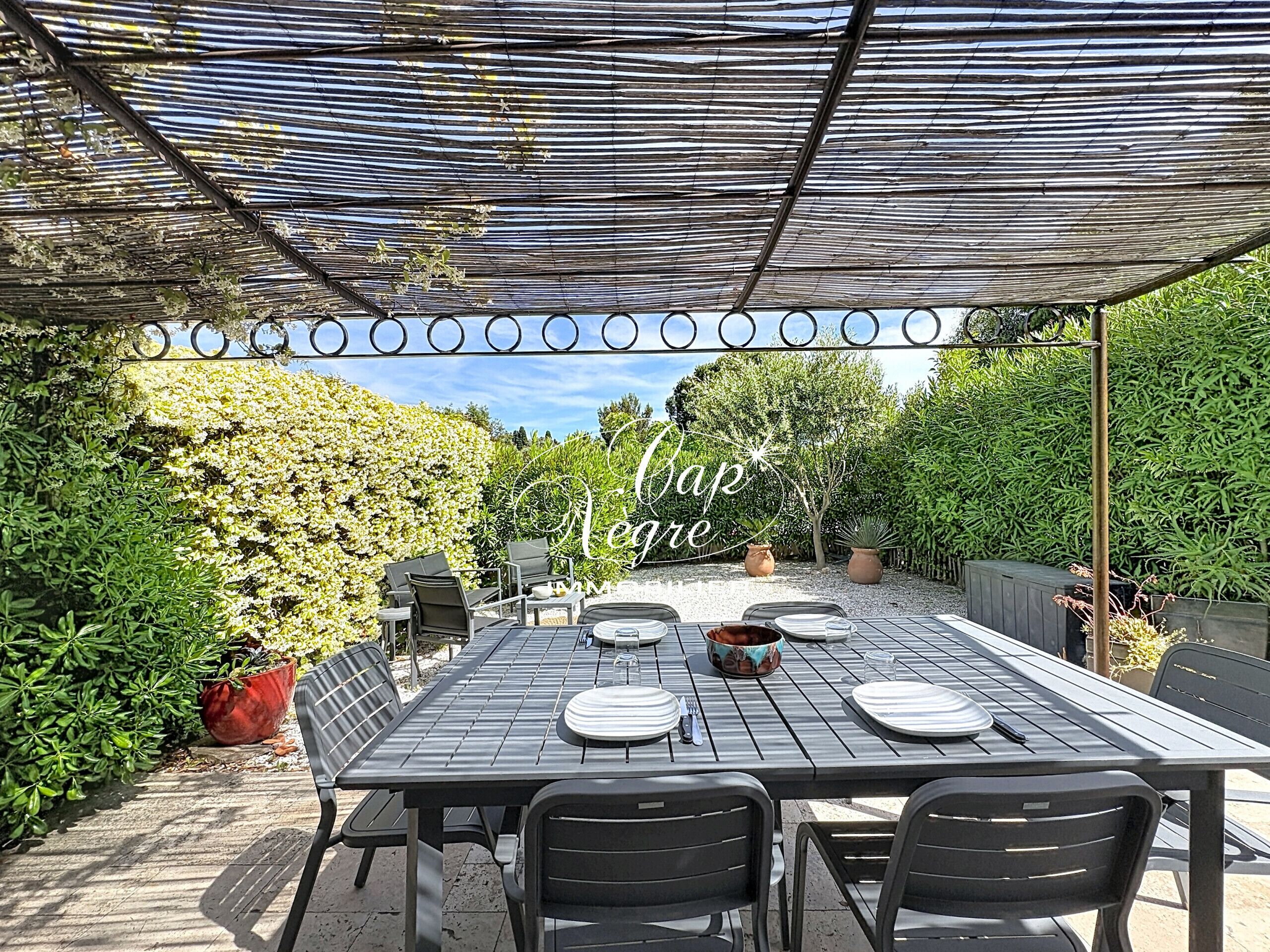 Une terrasse extérieure avec une table dressée pour le repas, entourée de verdure et surmontée d’une pergola.