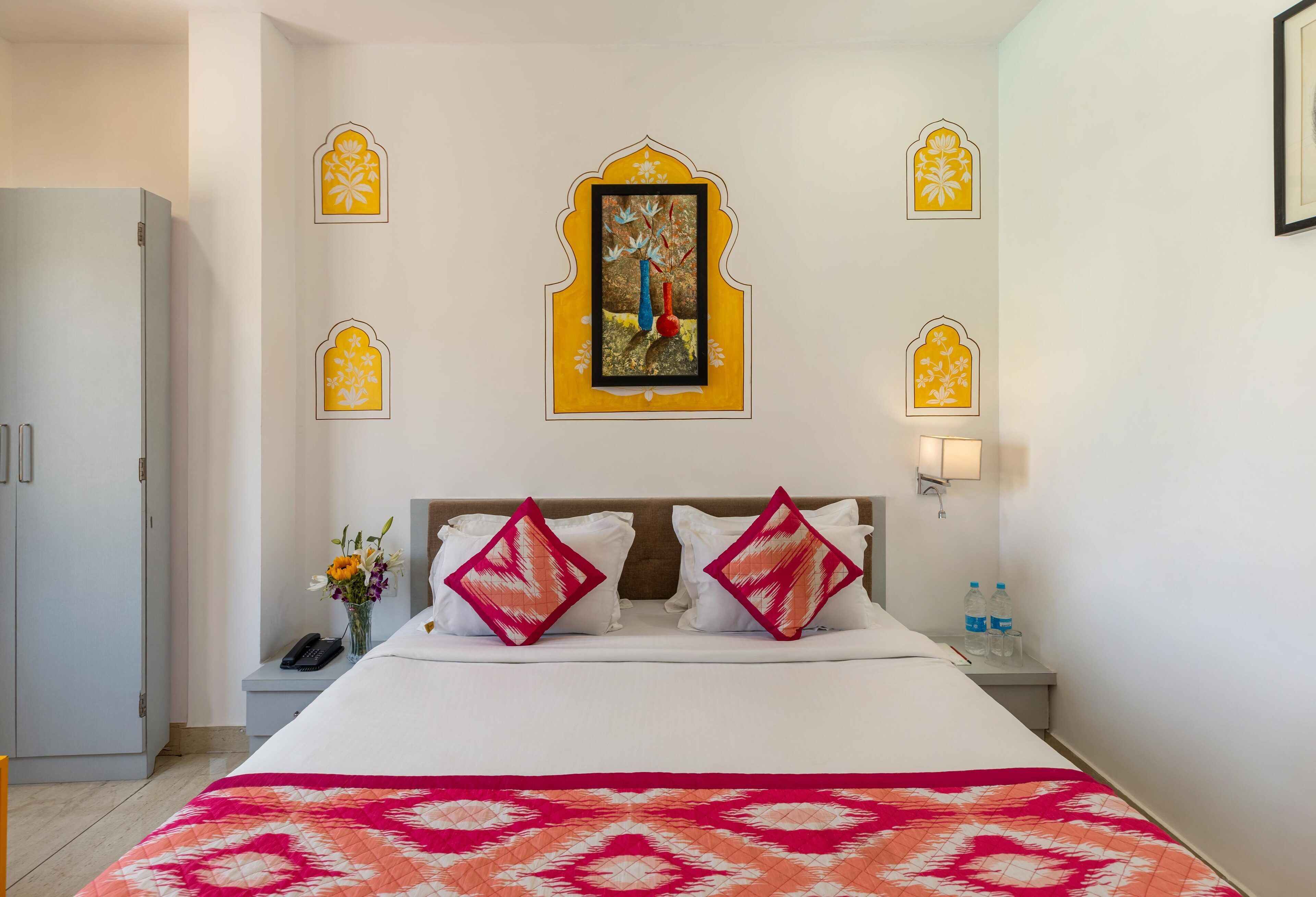 Une chambre d’hôtel avec un lit, des oreillers, une table de chevet, un téléphone, un vase avec des fleurs et un tableau accroché au mur.