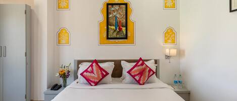 A hotel room with a bed, pillows, a nightstand, a phone, a vase with flowers, and a wall-mounted picture.