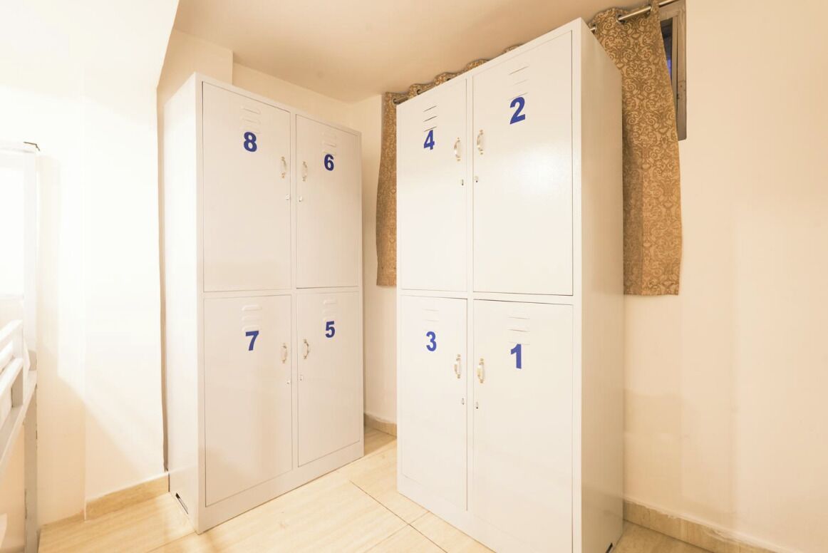 Una serie di armadietti bianchi, dal numero 1 all'8, in un angolo di una stanza con pavimento in legno e una finestra con tenda decorata.