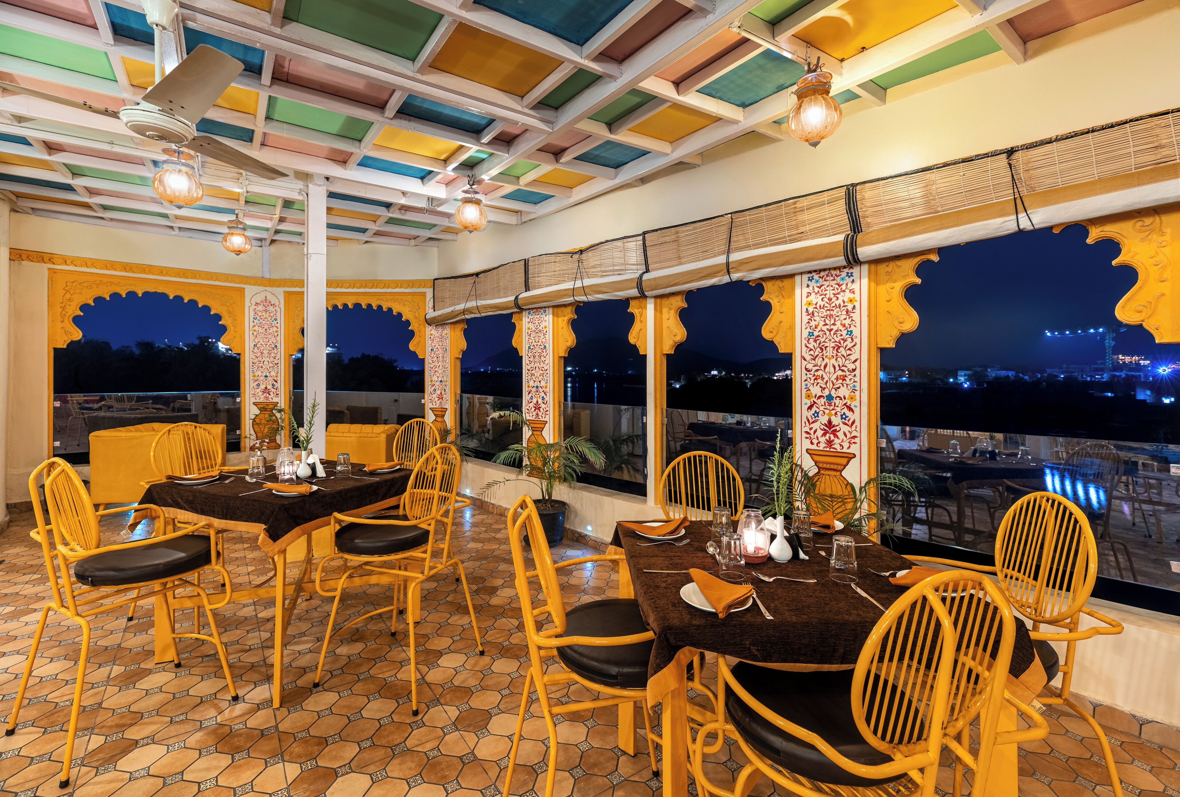 A dining area with a table set for four, surrounded by yellow chairs with black cushions. The room features a tiled floor and a ceiling with colorful panels.