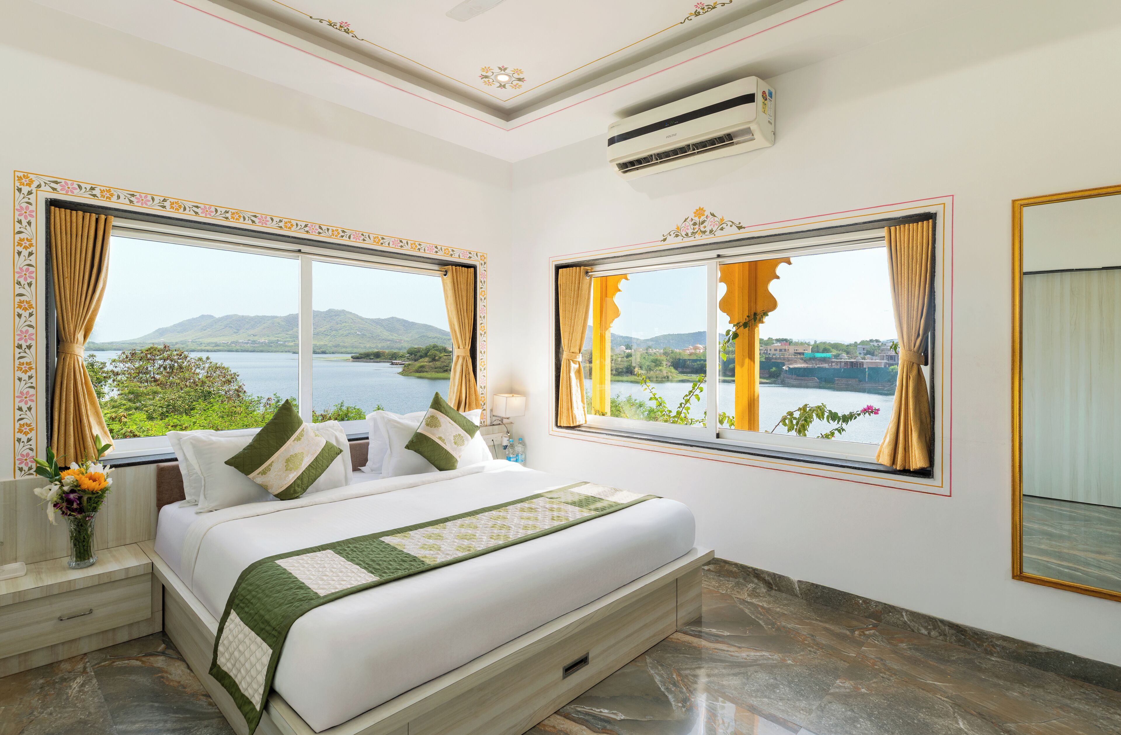 A bedroom with a large bed, two windows with curtains, a nightstand with a vase of flowers, and a view of a lake and mountains.