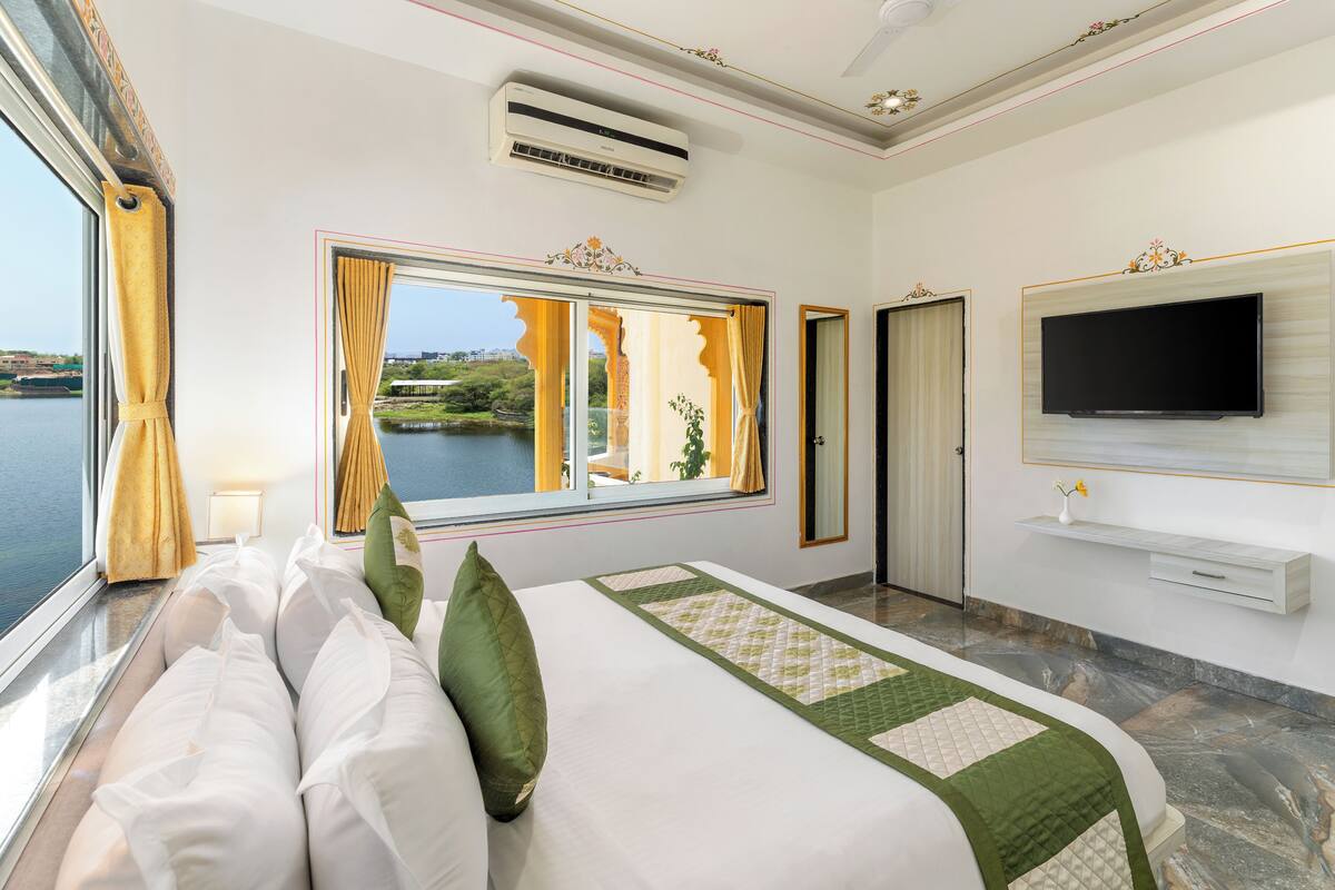 Una camera d'albergo con un letto, una televisione a parete, una finestra con tende e vista su un corpo d'acqua.