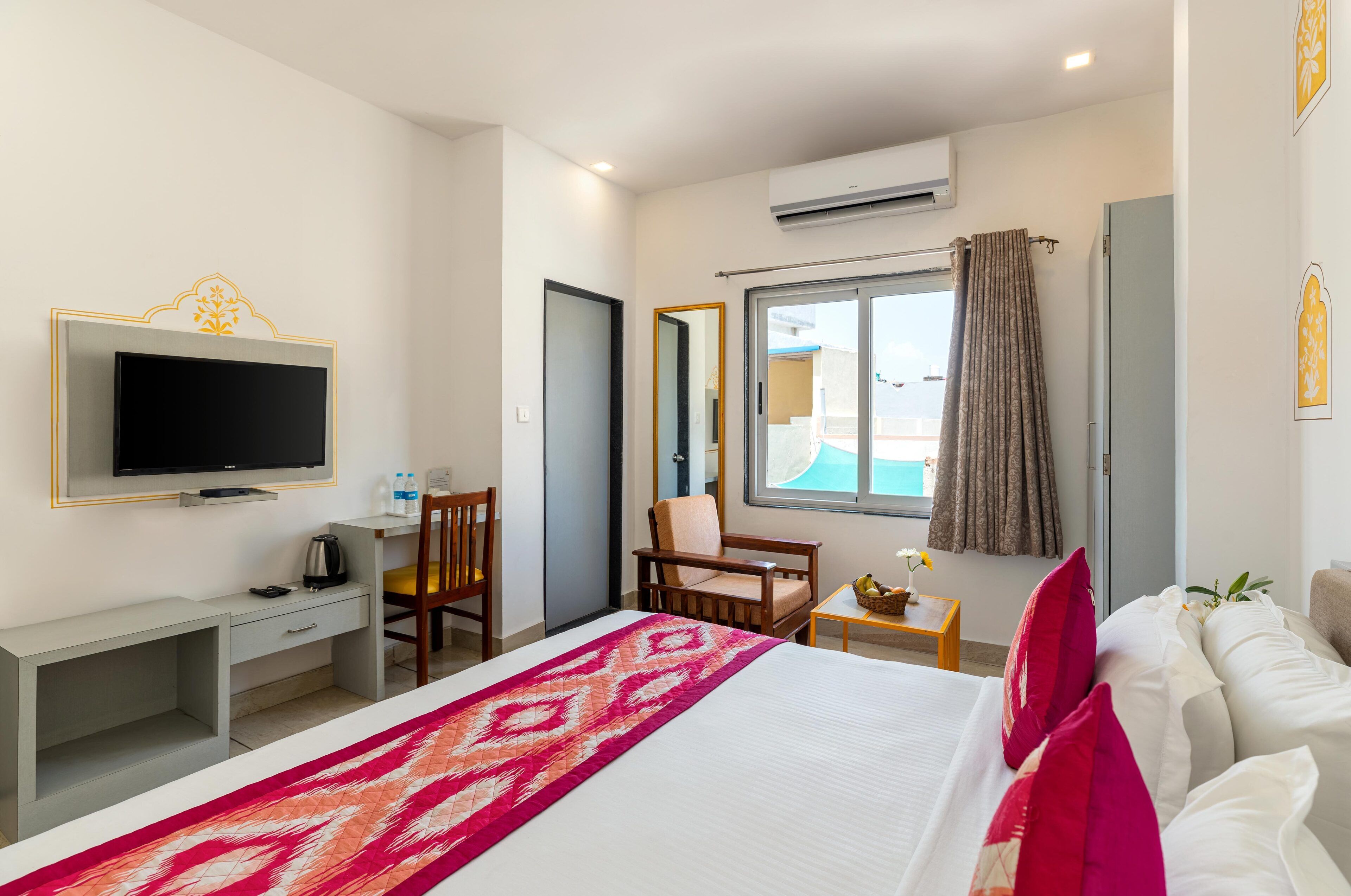 A hotel room with a bed, a desk, a chair, a television, a window with curtains, and an air conditioning unit.
