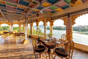 A restaurant with a view of a lake and mountains, featuring a table set for two with chairs, a ceiling with colorful panels, and a patterned floor. - Young Monk Hostel Udaipur - Jaydurg (Udaipur)