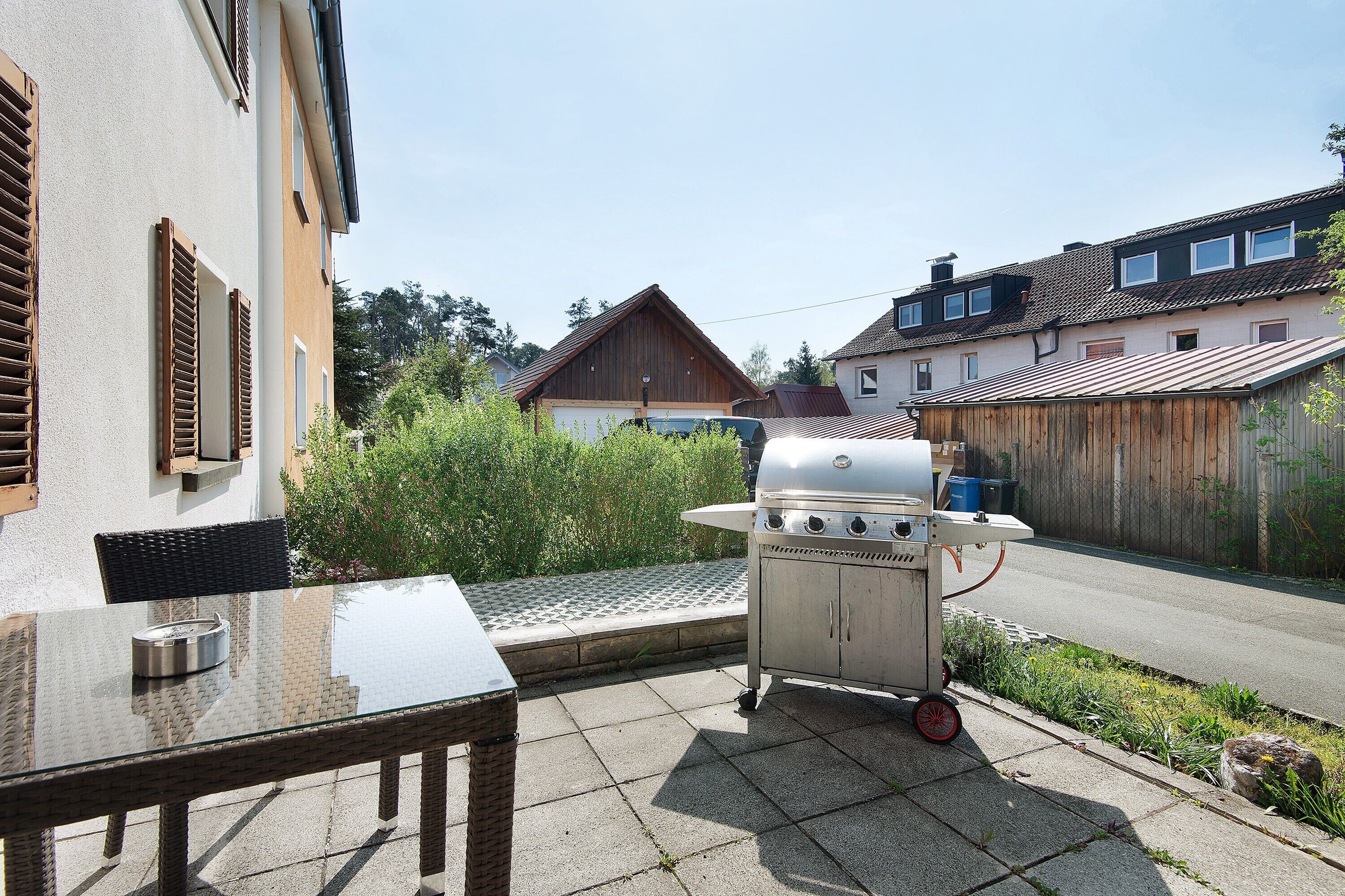 A patio with a glass table and chairs, a barbecue grill, and a view of a residential street.