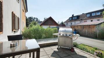A patio with a glass table and chairs, a barbecue grill, and a view of a residential street.
