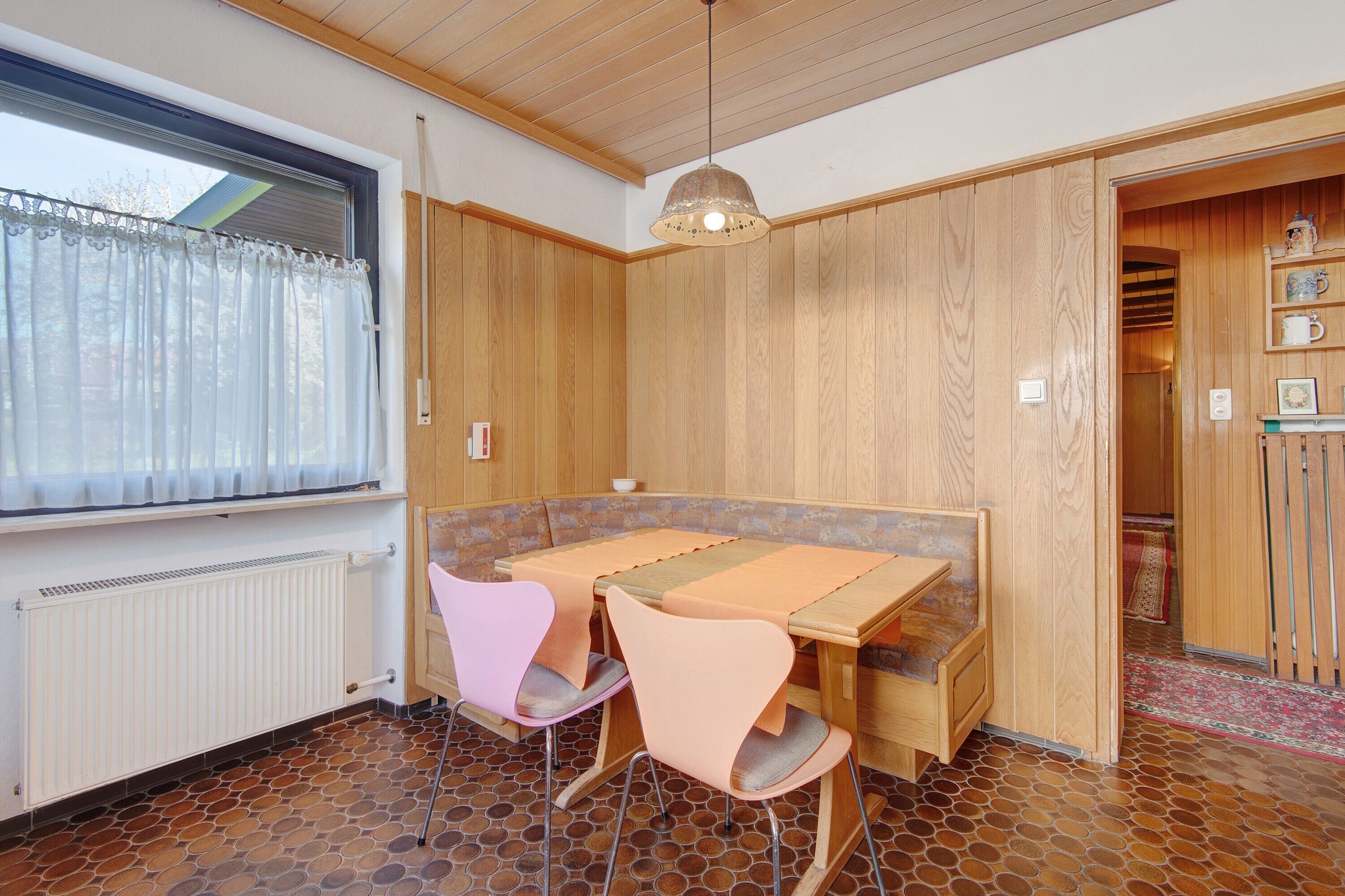 A dining area with a table and chairs, a window with curtains, a radiator, and a patterned floor.