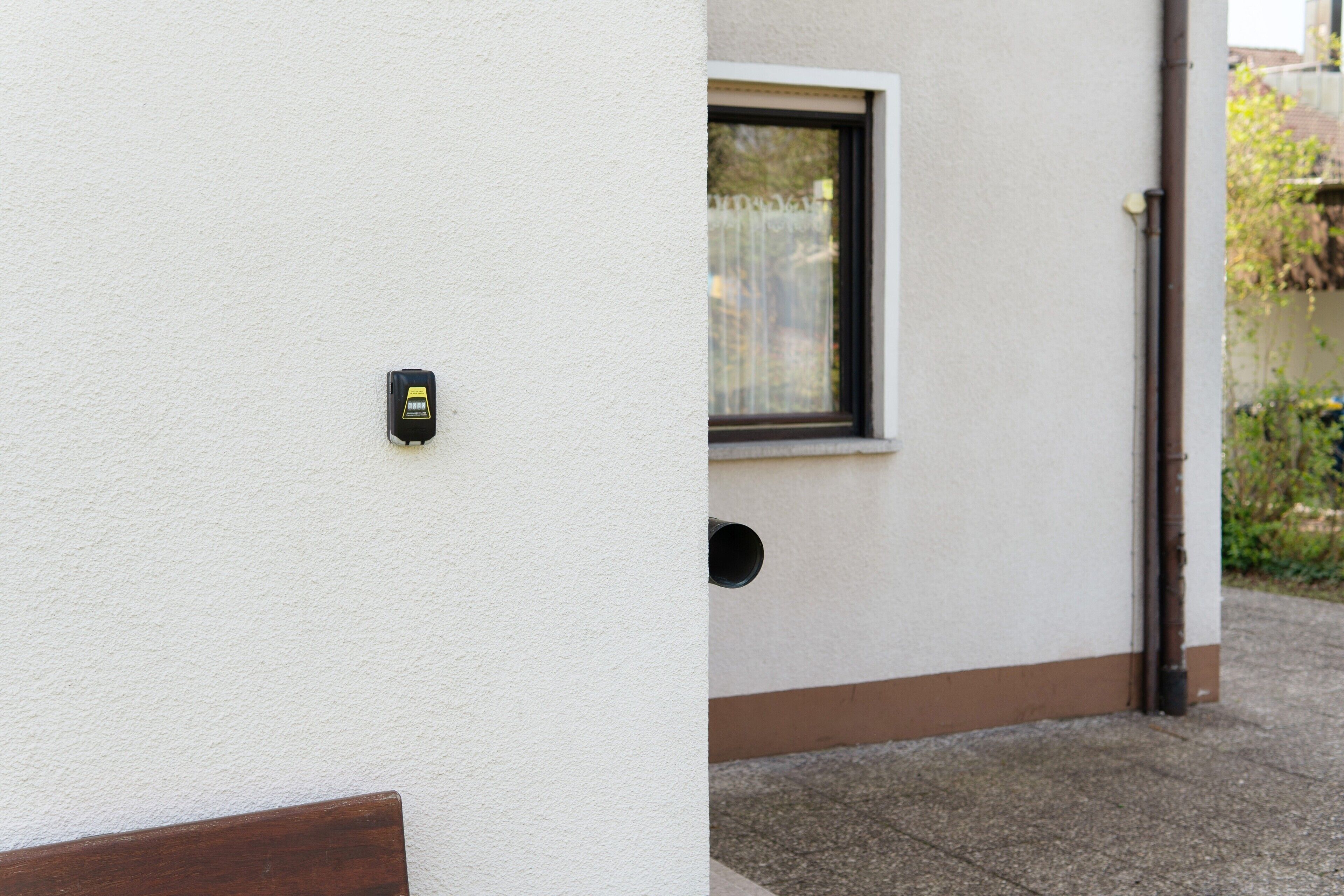 Un muro esterno bianco con un piccolo dispositivo nero fissato, una finestra con cornice marrone e un architrave marrone.