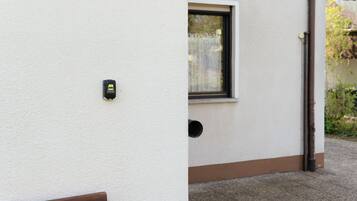 Un muro esterno bianco con un piccolo dispositivo nero fissato, una finestra con cornice marrone e un architrave marrone.