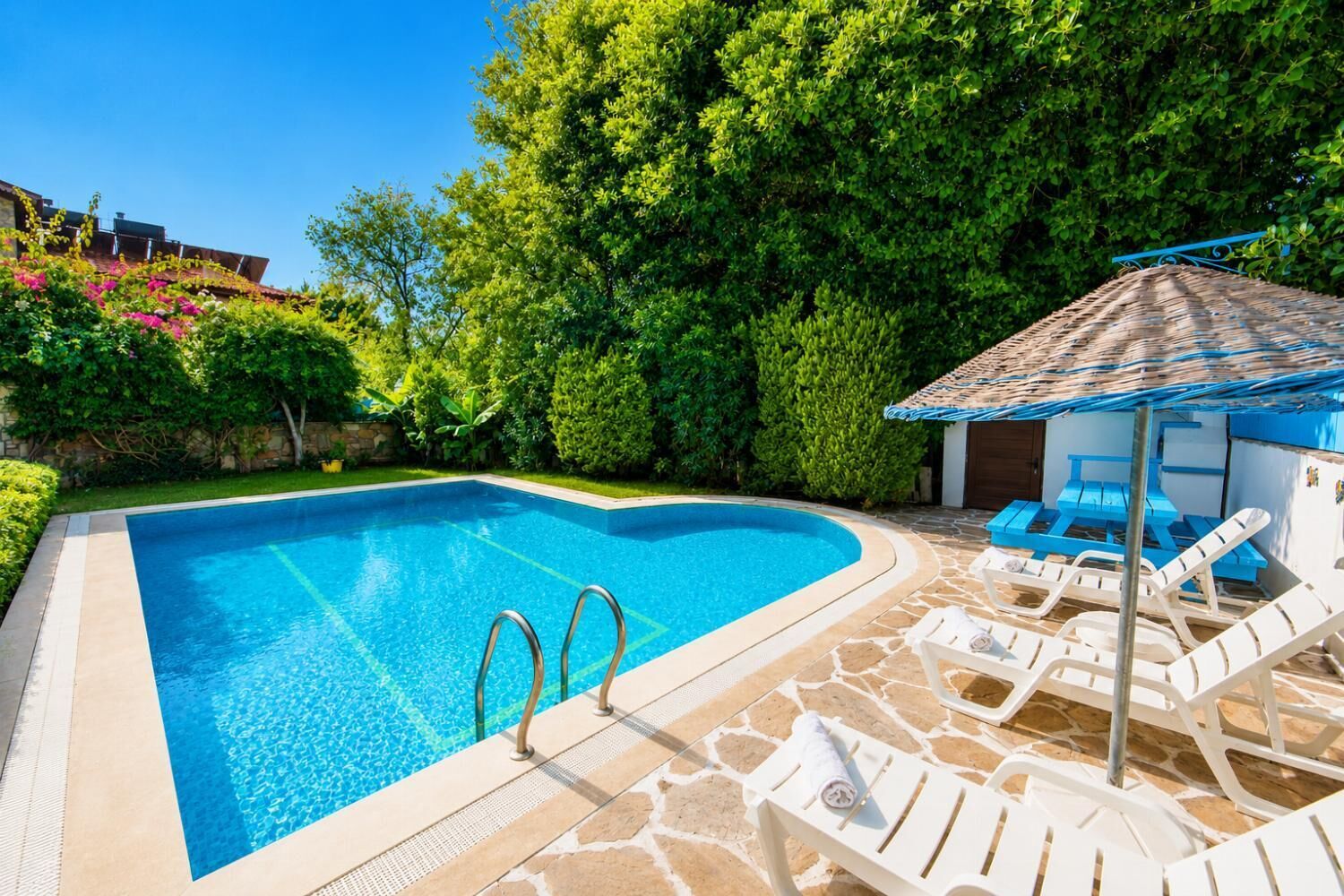 A swimming pool with a stone deck, lounge chairs, and a shaded area.