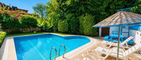 A swimming pool with a stone deck, lounge chairs, and a shaded area.