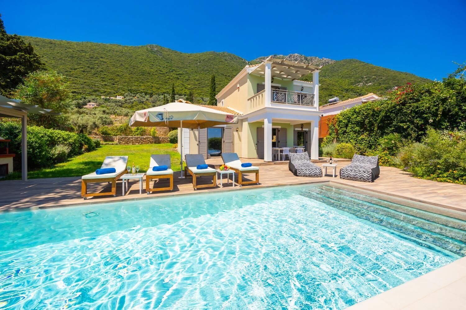 A pool area with lounge chairs and umbrellas, a two-story building with a balcony, and a mountainous backdrop.