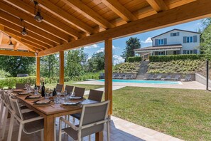 Un espace extérieur aménagé pour les repas, avec une pergola en bois, une piscine et une maison en arrière-plan.