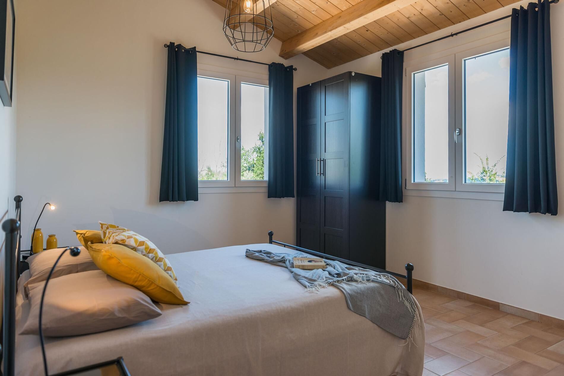 Ein Schlafzimmer mit Bett, Kleiderschrank, zwei Fenstern mit Vorhängen, einer Lampe und Blick auf Grünfläche.
