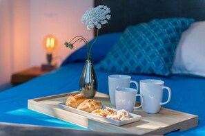 Un plateau de petit-déjeuner avec des viennoiseries et des tasses, posé sur un lit avec une literie bleue et un vase rempli de fleurs.
