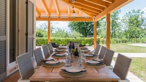 Ein überdachter Essbereich im Freien mit einem Holztisch, gedeckt für sechs Personen, umgeben von Stühlen und Blick auf einen Garten.