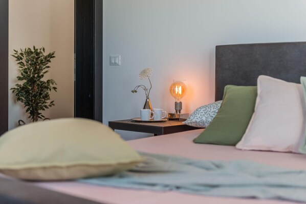 A neatly made bed with pillows, a bedside table with a lamp, a plant, and a cup.