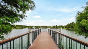 A wooden bridge with metal railings leading over a calm body of water, surrounded by lush greenery.