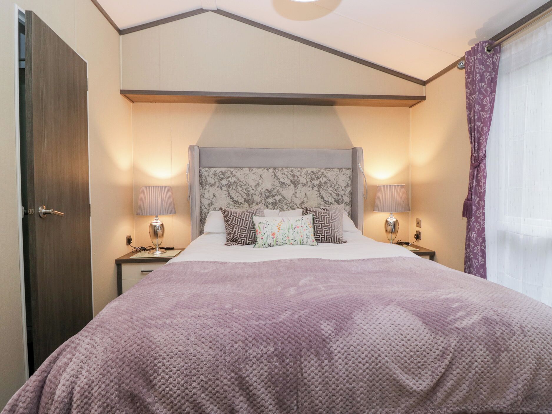 Un dormitorio ordenado con una cama grande, dos mesitas de noche con lámparas, una puerta de madera y una ventana con cortinas.