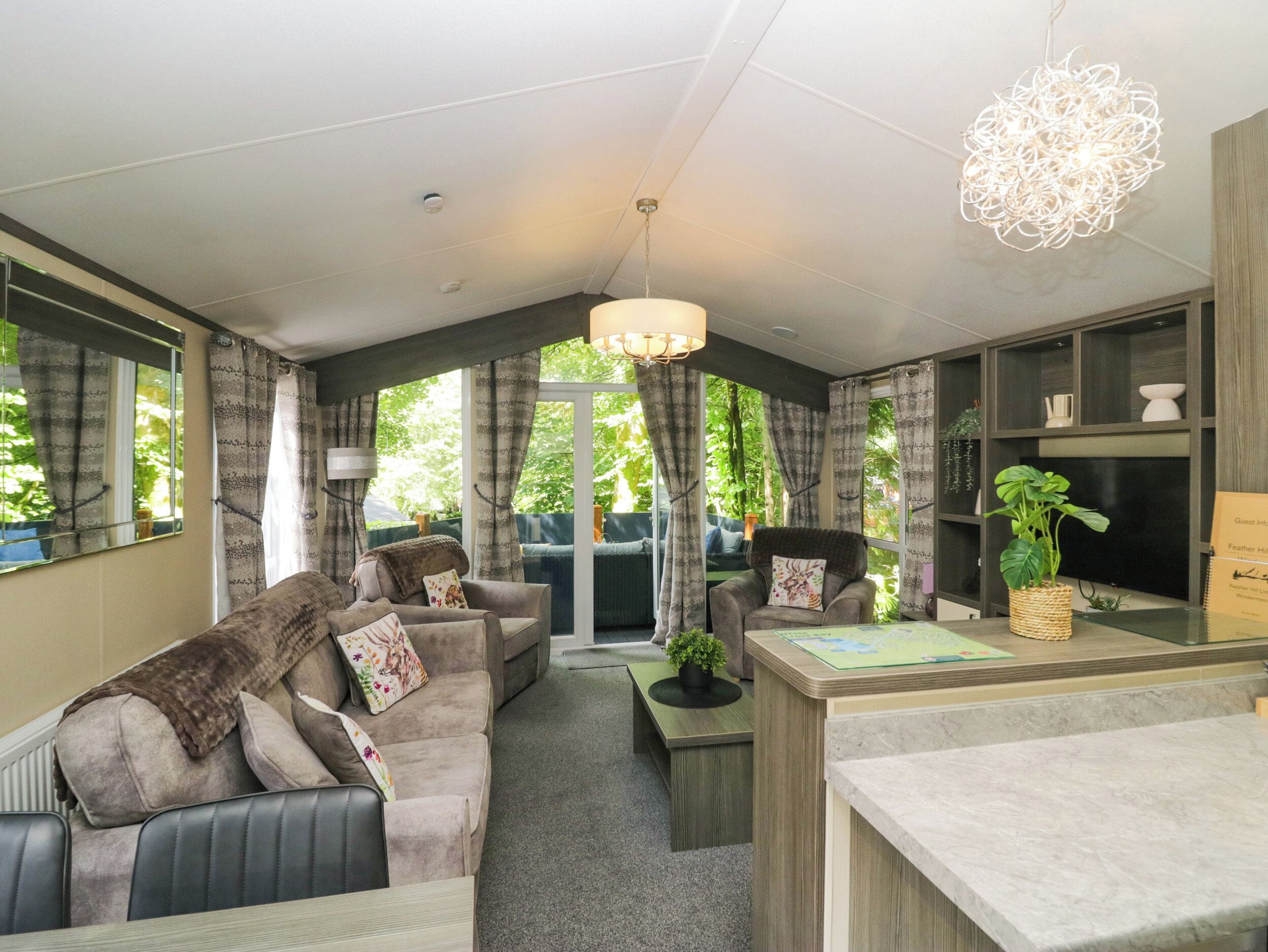 A cozy living area with a sofa, armchairs, a coffee table, and a dining table. There is a large window with curtains, a hanging light fixture, and a bookshelf.