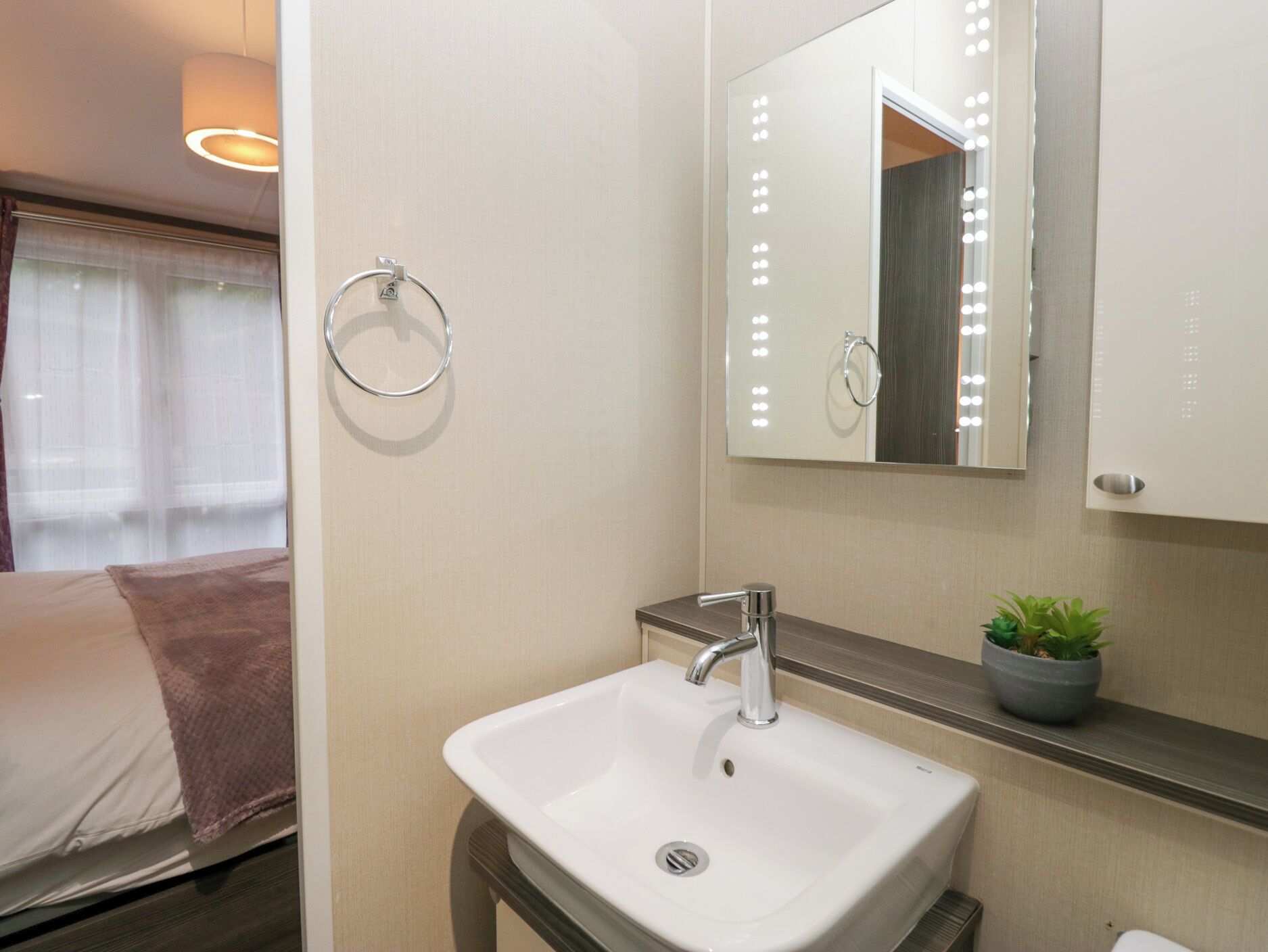 Un baño moderno con un lavamanos blanco, un espejo con iluminación, un sujetarrepisas para toallas y una pequeña planta en maceta.