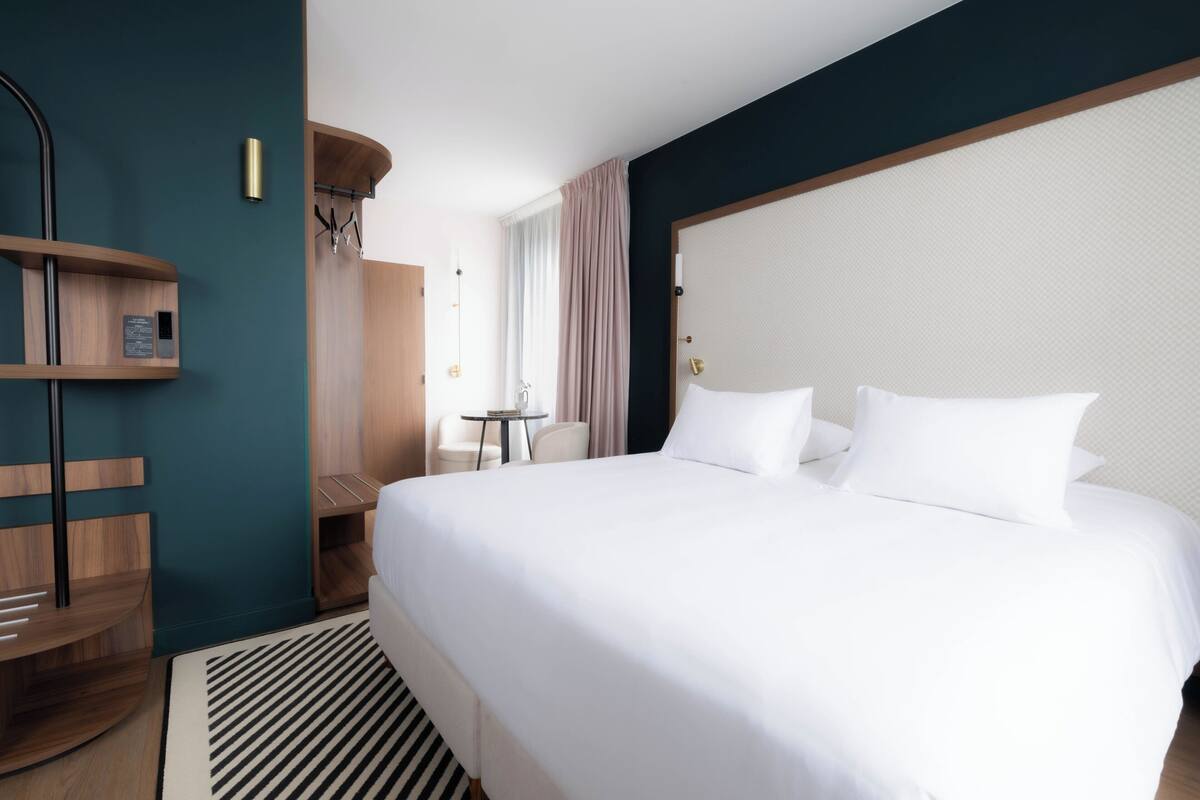 A hotel room with a large bed, a small table, a chair, a wooden shelf, and a wall-mounted lamp.