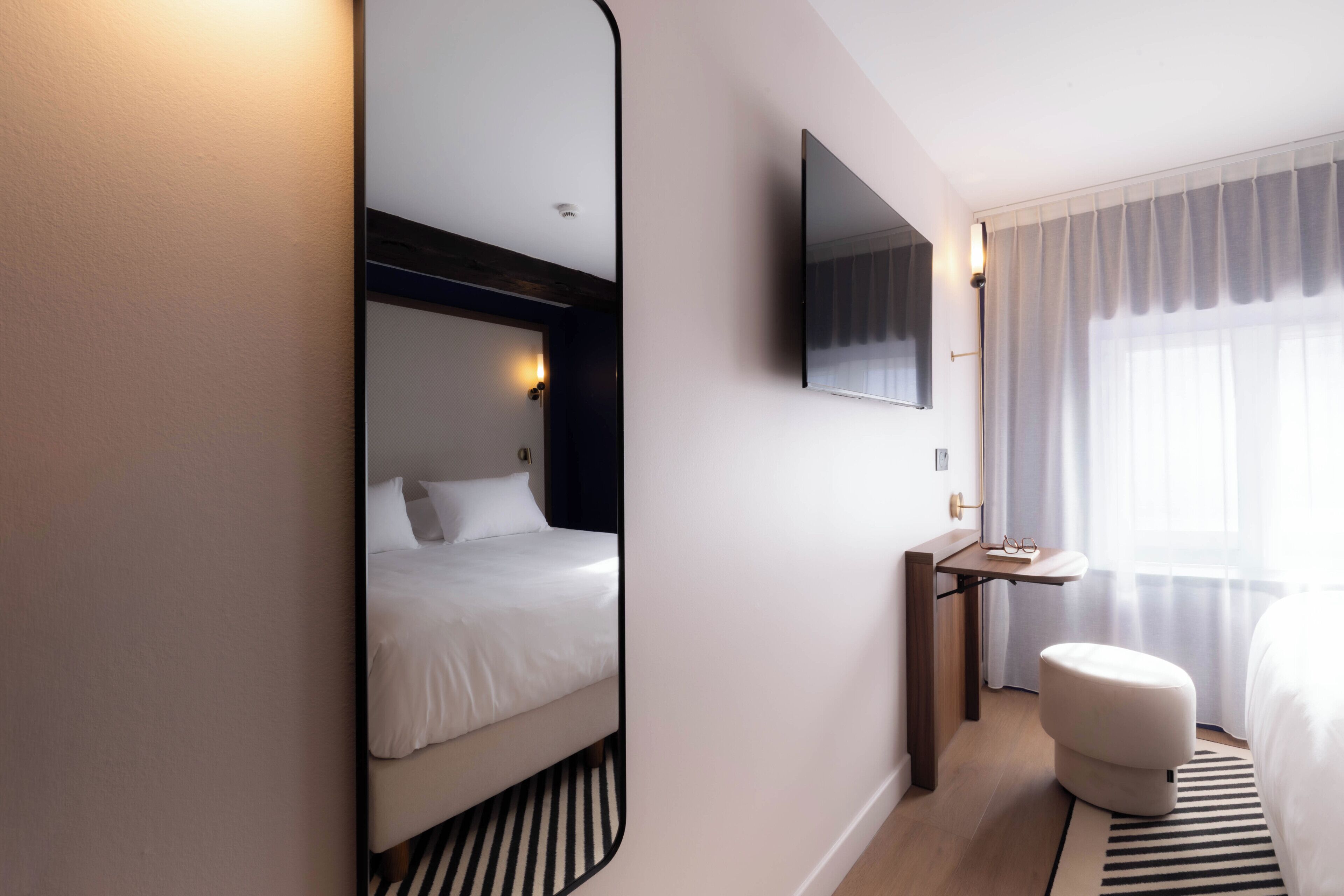 Quarto de hotel moderno com cama, mesa de cabeceira, espelho, TV de tela plana, um banquinho pequeno e janela com cortinas.
