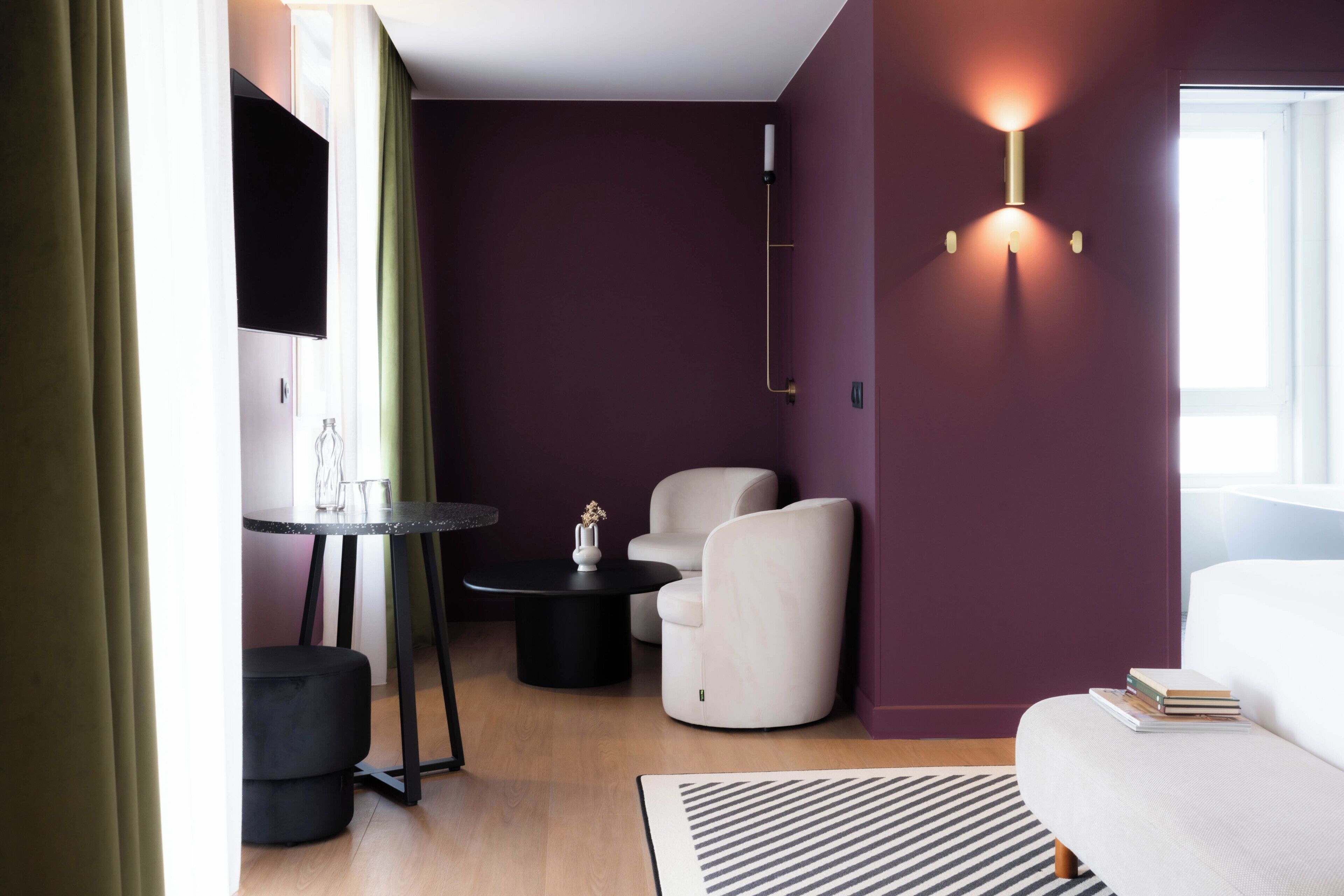 A modern hotel room with a sofa, a round table, a chair, a flat-screen TV, and a wall-mounted light fixture.