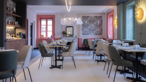 Uma sala de jantar moderna com várias mesas e cadeiras, uma cozinha ao fundo e iluminação decorativa nas paredes.