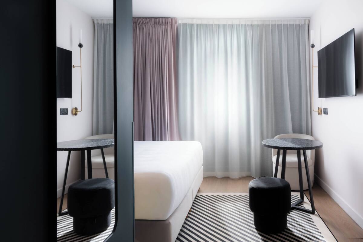 A modern hotel room with a bed, two side tables, a TV, and a large window with curtains.