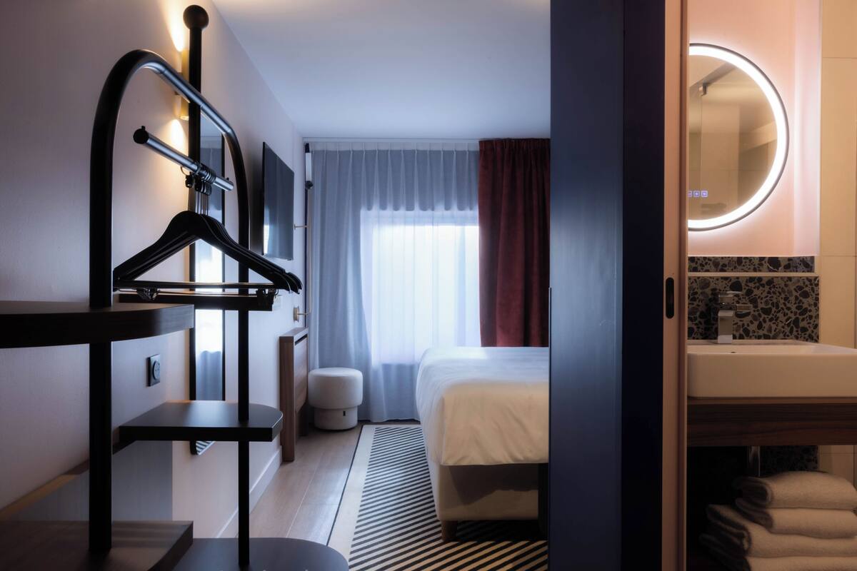A modern hotel room with a bed, a hanging coat rack, a bedside table, a round mirror, and a bathroom with a sink and towels.