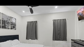 Quarto moderno com cama grande, duas janelas com cortinas, ventilador de teto, televisão montada e cômoda.