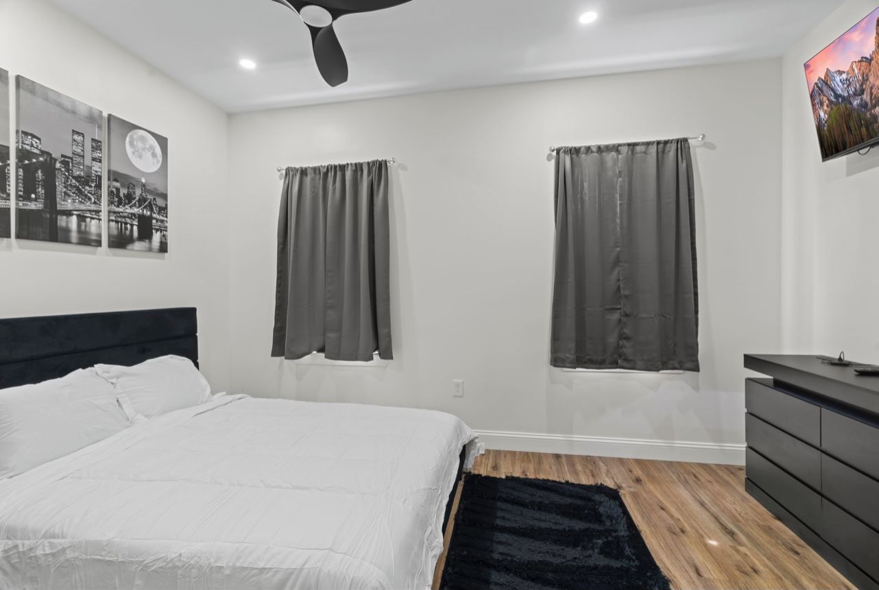Ein modernes Schlafzimmer mit einem großen Bett, zwei mit Vorhängen versehenen Fenstern, einem Deckenventilator, einem wandmontierten Fernseher und einer Kommode.