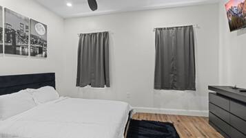 Ein modernes Schlafzimmer mit einem großen Bett, zwei mit Vorhängen versehenen Fenstern, einem Deckenventilator, einem wandmontierten Fernseher und einer Kommode.
